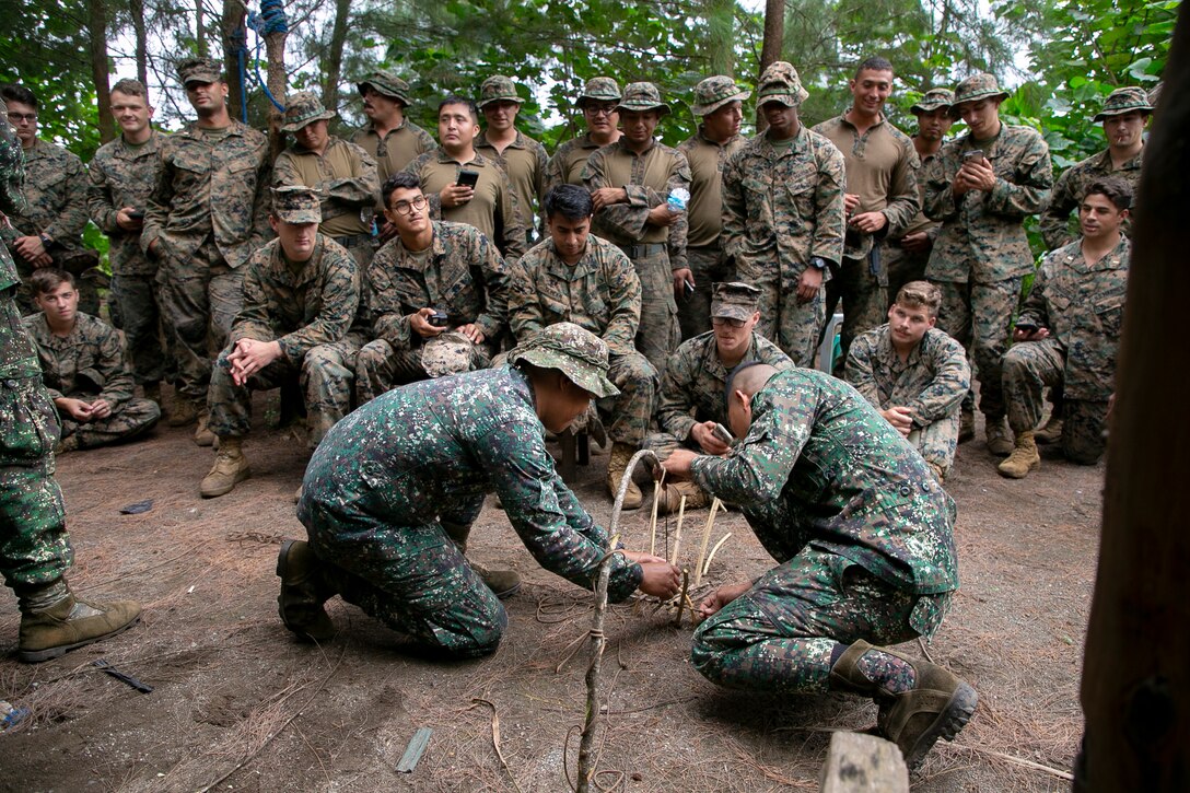 More than a dozen uniformed military personnel stand near each other and watch two other personnel use sticks to build an apparatus on the ground.