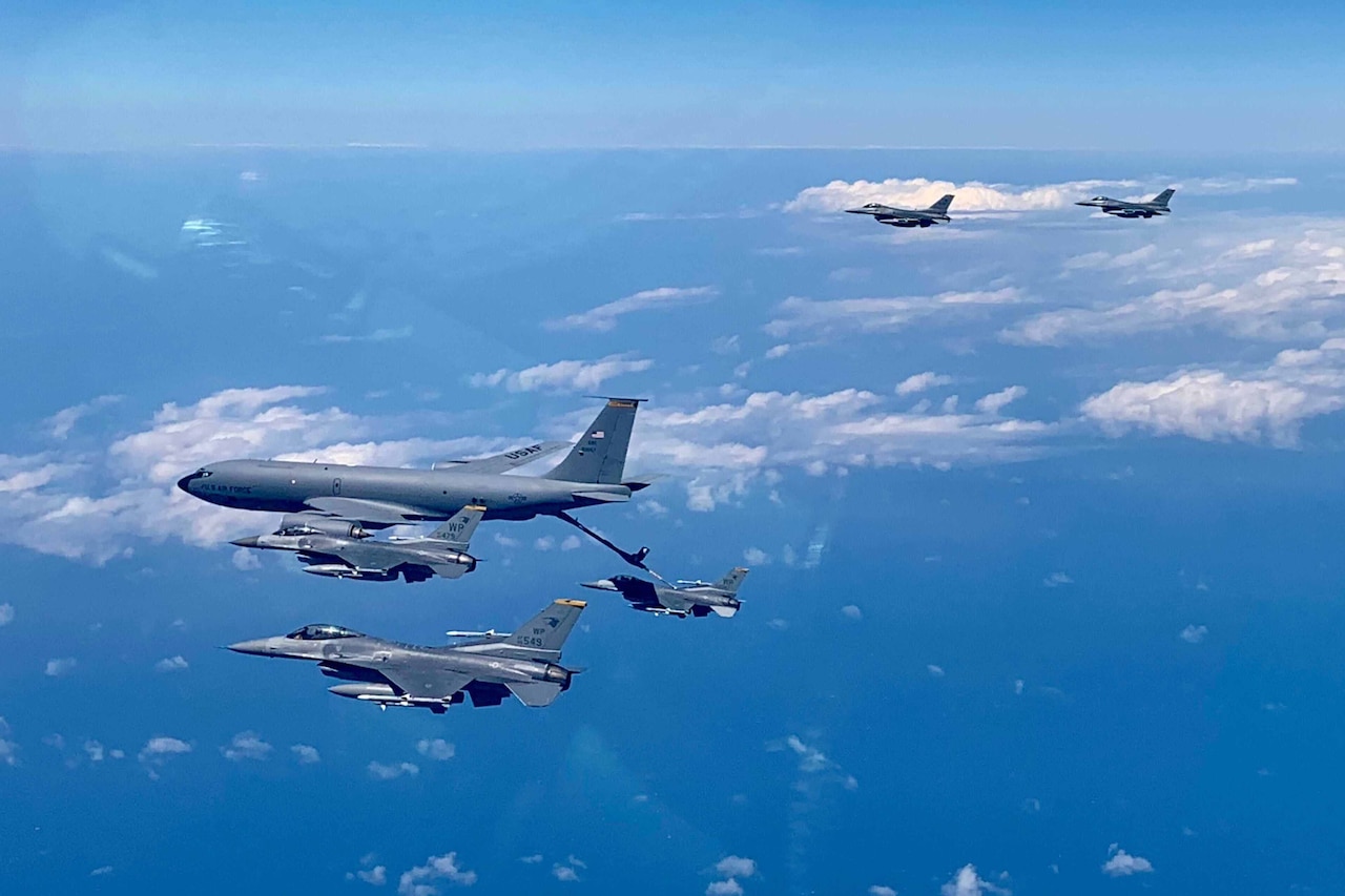 Several military fighter jets fly near a larger tanker aircraft. One is tethered to the tanker.