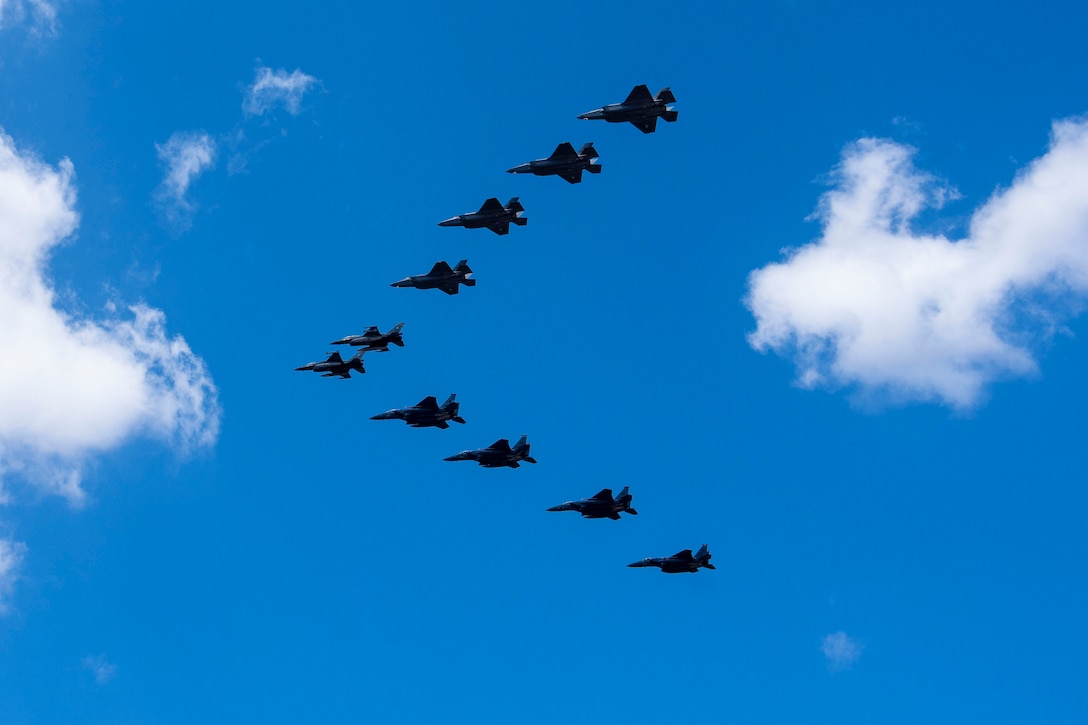 Ten aircraft fly in a v formation against a blue sky with scant clouds.