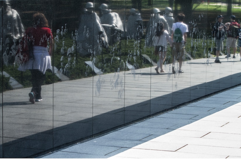 A black, granite wall displays engravings of military members, and its surface reflects visitors passing by.