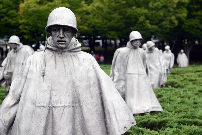 A stainless steel statue of a man in a military-style helmet and rain poncho is positioned in a landscaped area near similar statues.