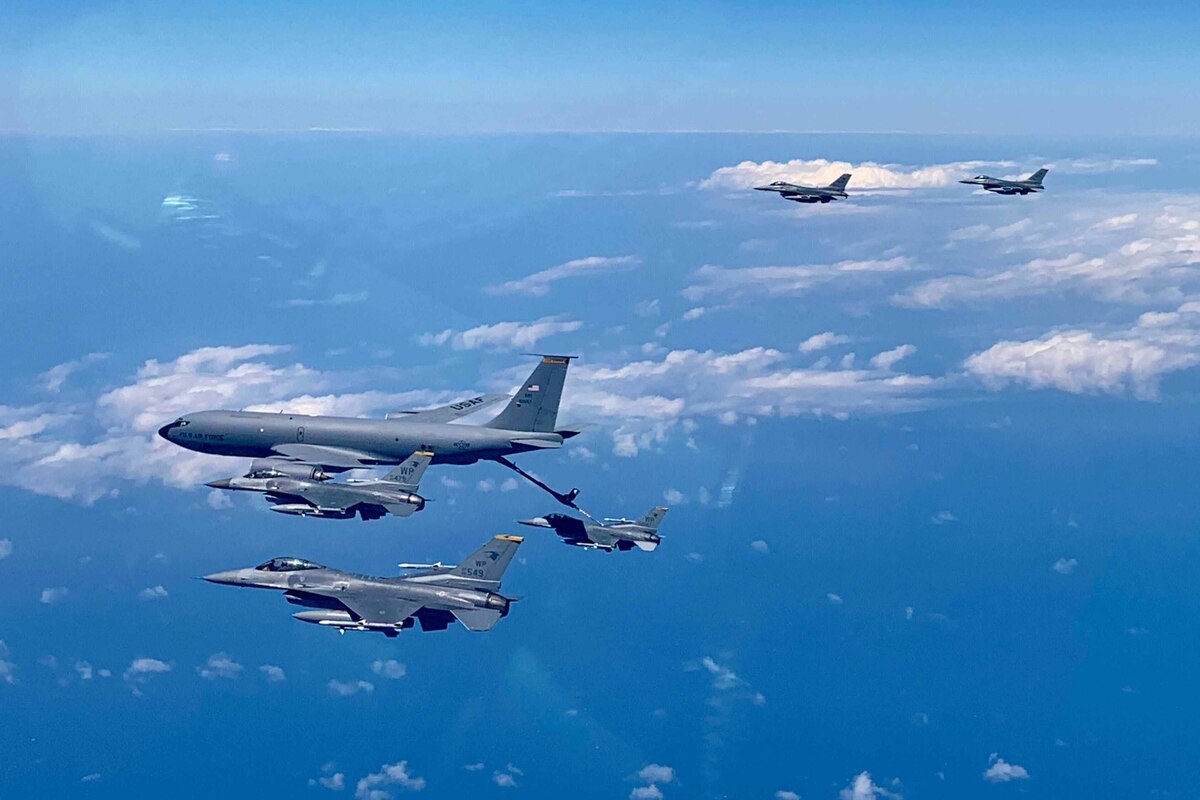 Several military fighter jets fly near a larger tanker aircraft. One is tethered to the tanker.