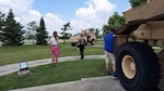 A woman in a white sleeveless blouse and pink flowered skirt interviews a woman in Army dress blues in a park setting with military equipment. A cameraman dressed in a blue shirt and tan shorts holds a camera on a tripod in front of a large tan military vehicle on static display. The woman in the uniform stands on a sidewalk and the woman in the white blouse and pink flowered skirt stands on grass. A light for lighting the military vehicle sits behind the woman in the pink skirt. The woman in uniform has the dress pants with a gold border version of the uniform and has many ribbons and medals on the uniform coat.