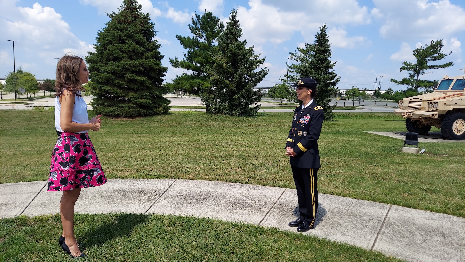 A woman in a white blouse with no sleeves and a pink flowered skirt and high heels stands on grass interviewing a woman in Army dress blues (black uniform with gold stripes, ribbons and medals and a black gold trimmed hat). She wears the dress pants with gold trim and black shiny shoes. She stands on a white sidewalk in a park with trees and military equipment. Part of a brown military vehicle can be seen behind her.