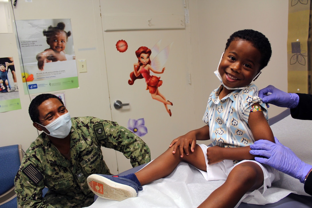 A little boy sits on an examination table and smiles at the camera while a person with glove-clad hands reveals a small adhesive bandage on his upper arm.