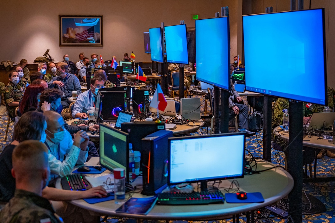 A room of service members and other personnel sit at tables with an abundance of computers in front of several large display monitors.