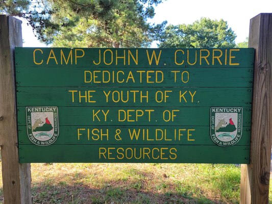 A green sign with the words Camp John W. Currie dedicated to the youth of Ky. Ky Dept. of Wildlife Resources