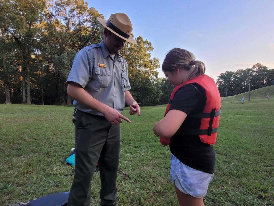 Ranger Ben Clark stands in a field and instructs a young female camper on the proper way to fit a life jacket.  (USACE Photo by WES DAVENPORT)
