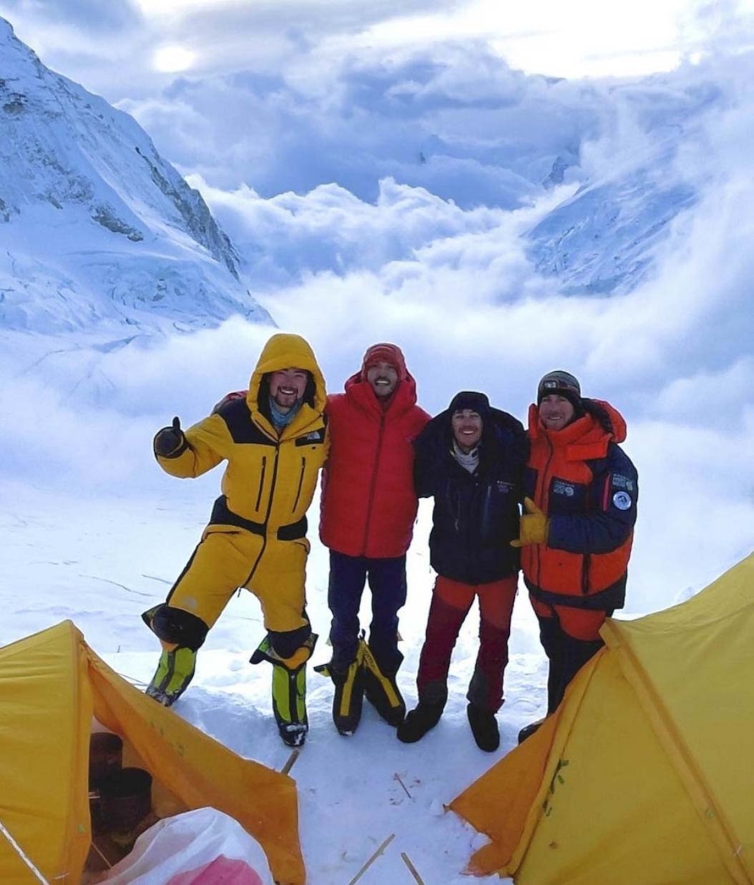 Four men dressed in winter gear pose in front of a cloud and snow-covered mountain.