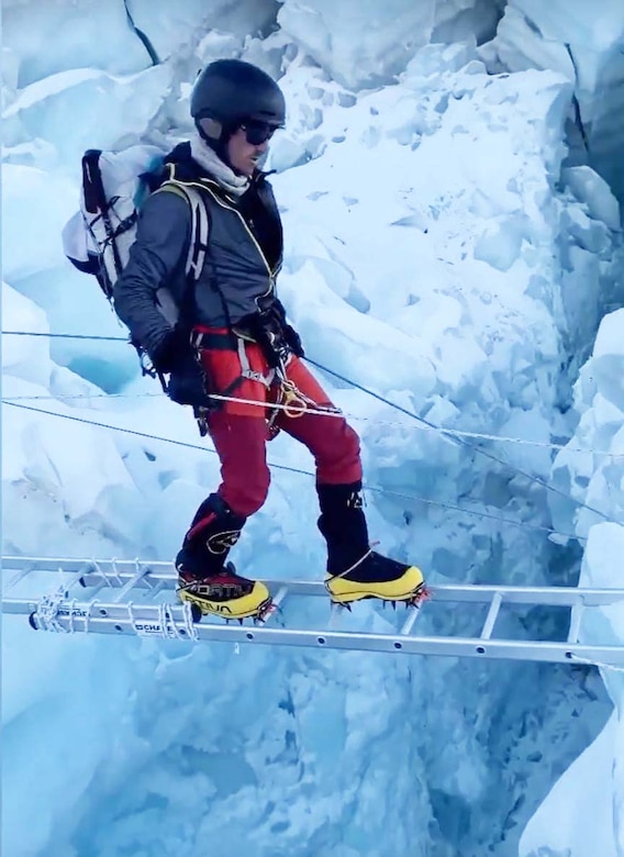 A man dressed in cold weather gear, walks across a ladder used as a bridge on a snow-covered mountain.