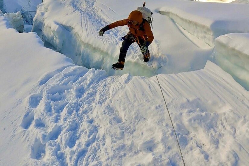 A man dressed in cold weather gear, jumps across a large crack in the snow.
