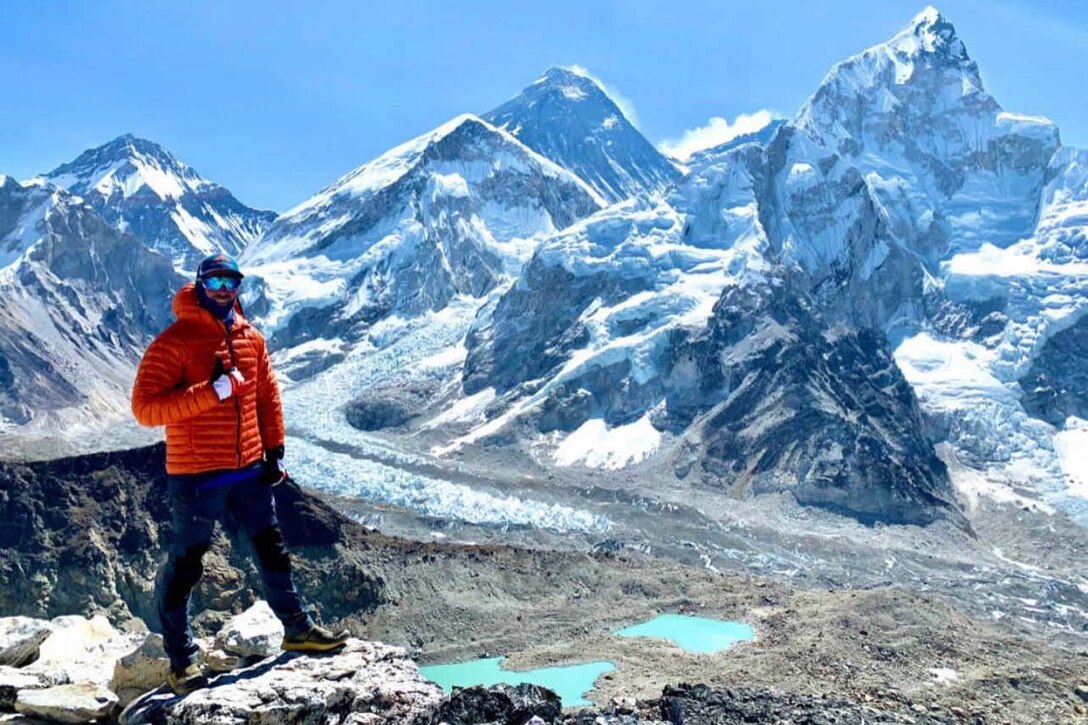 A man dressed in cold weather gear stands in front of tall mountains.