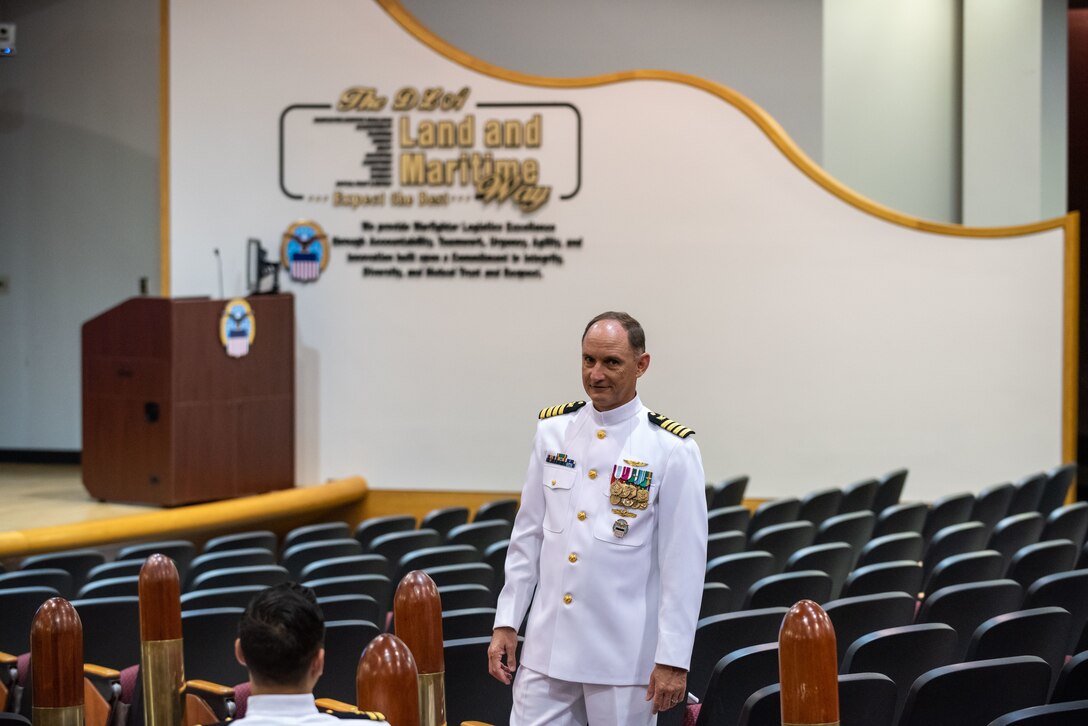 A man in Navy dress whites with gold trim on hsi shoulders and sleeves stands in a auditorium with large bullet style decorations lining the aisle.