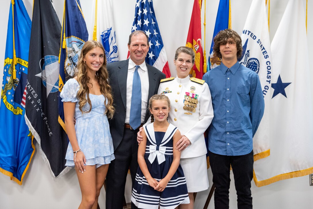 (From left) a woman in a short blue prairie style dress, a man in a blue suit and blue tie, a woman in Navy dress whites with gold trim and a skirt, and a man in a blue dress shirt and black pants pose in front of a grouping of military flags. In front of the woman in dress whites is a girl in a blue and white striped dress.