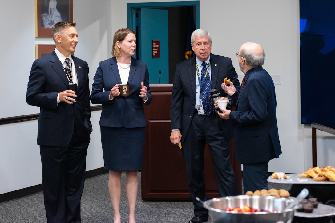 A man with a buzz cut wearing a black suit stands next to a woman in a dark blue suit and white blouse who stands next to a man with gray hair in a blue suit who chats with a balding man with a bears in a dark suit. They are surrounded by food on a table and are in a conference room.
