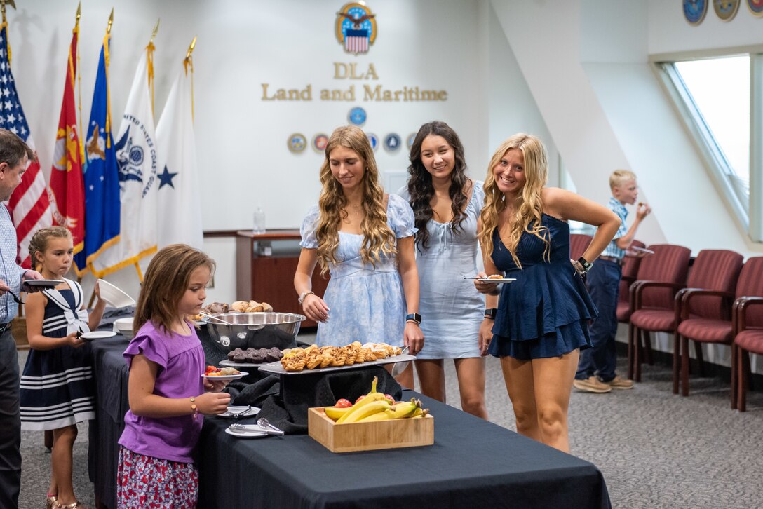 Several young adults and children in a conference room smile as they grab food from a table with a black tablecloth.
