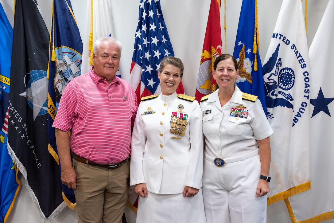 A bald man in a pink shirt stands next to a woman in Navy dress whites with gold trim, ribbons and medals. The woman is in a white skirt with white shoes. She is flanked by another woman in Navy dress whites (pants version). They stand next to a grouping of military flags against a wall in a conference room.