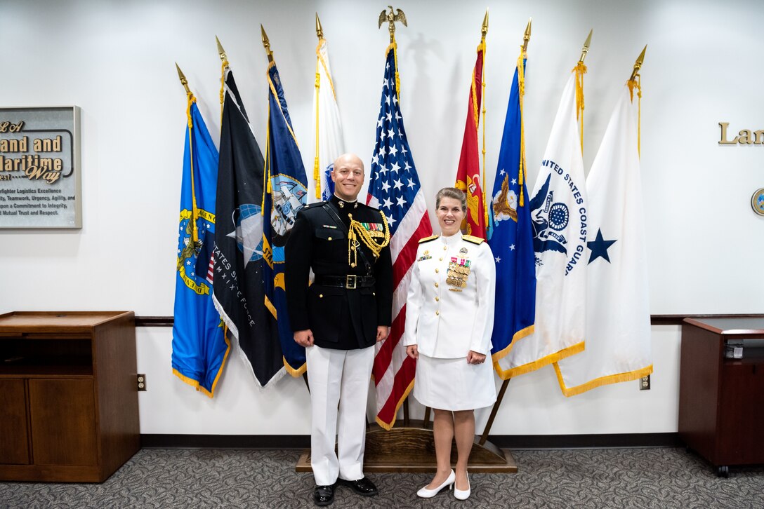 A bald man in a Marine Corps dress uniform with ribbons and gold trim (black service coat and white pants) stands next to a woman in Navy dress whites with gold trim, ribbons and medals. The woman is in a white skirt with white shoes. They stand next to a grouping of military flags against a wall in a conference room.