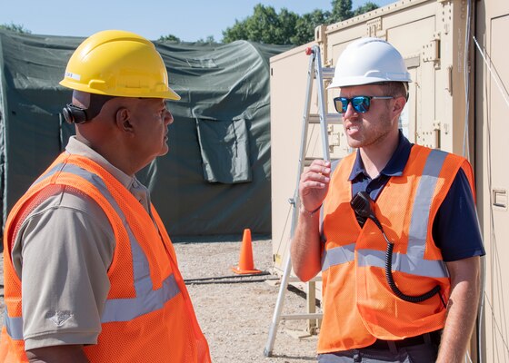 Two men in hardhats discuss operations in front of portable structures.