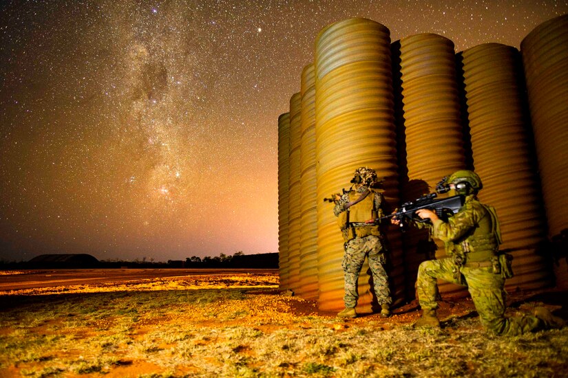 Two soldiers, one standing and one kneeling, hold weapons at night.