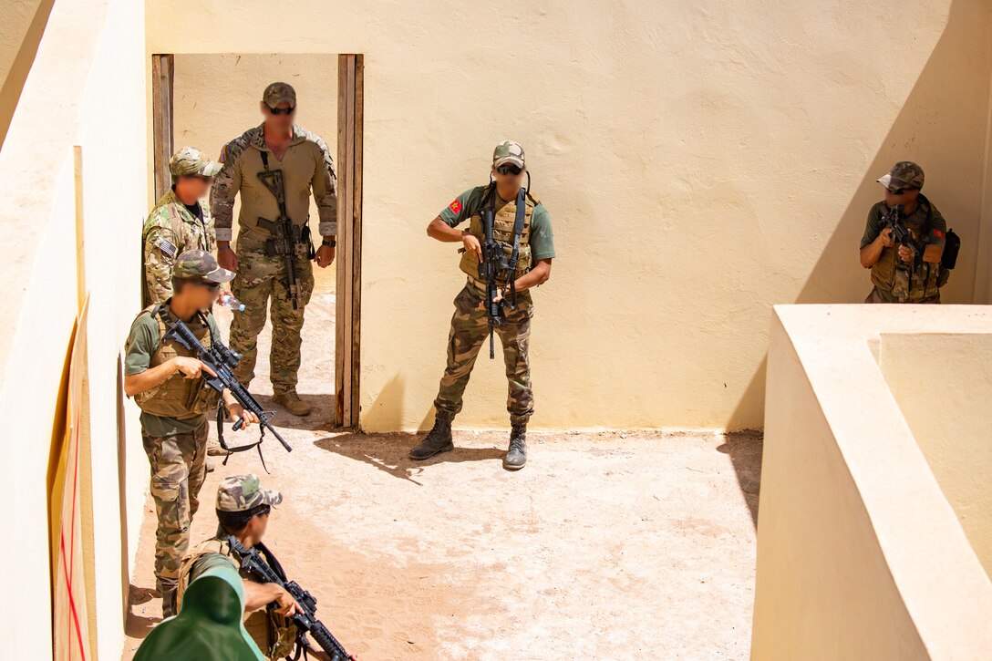 Military personnel hold weapons and stand around in a small room.