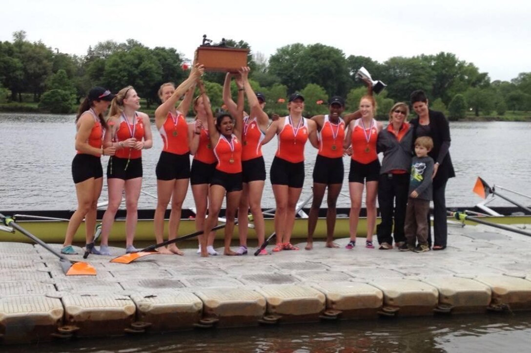 Several female rowers hold up a trophy on a dock as they pose for a photo.