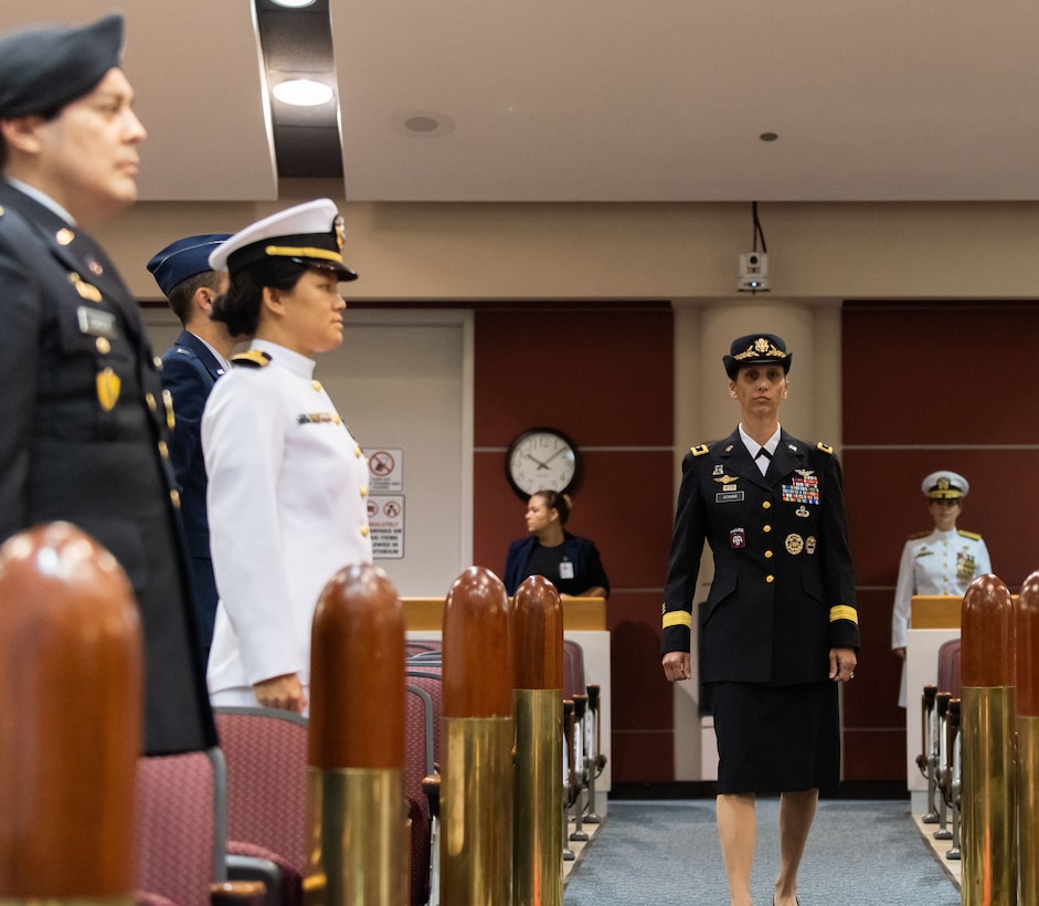 A woman in Army dress blues which is actually a black color walks down the aisle of an auditorium while a woman in Navy dress whites stands behind her. Military members in Navy and Army dress uniforms flank the women in the back with reproductions of bullets on either side.
