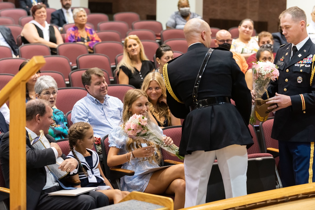 A man in a US Marine Corps uniform with a black coat with gold trim and white pants gives a gift to family members of Commander Fabry who are seated in a auditorium. He gives what looks like to be a box to Fabry's husband who is dressed in a grey suit with blue tie.