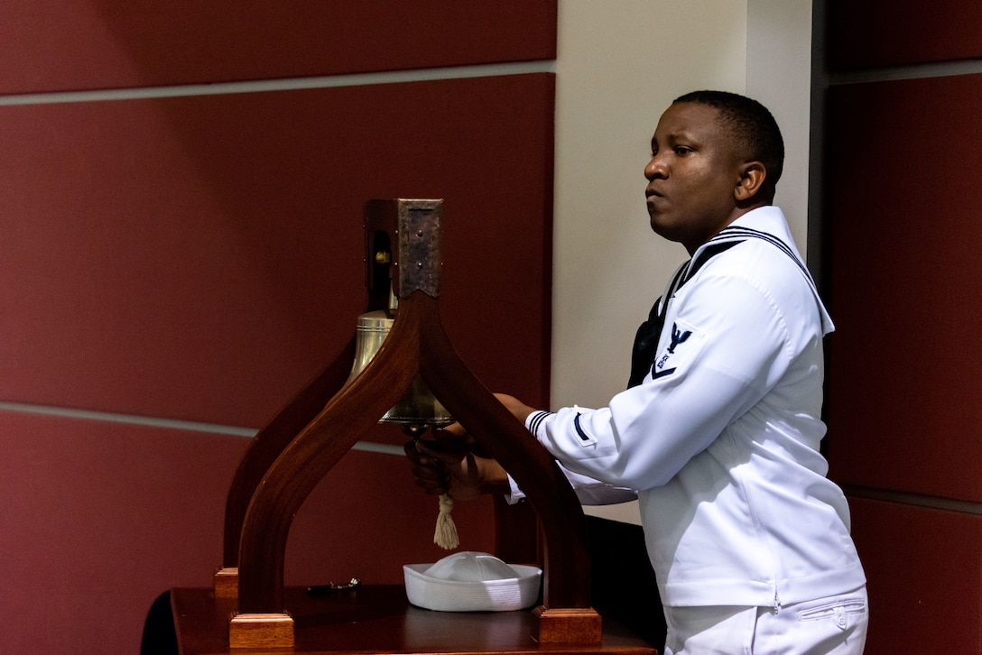 A man in a Navy enlisted uniform with a black tie and a round Navy cap rings a bell signifying The Watch.
