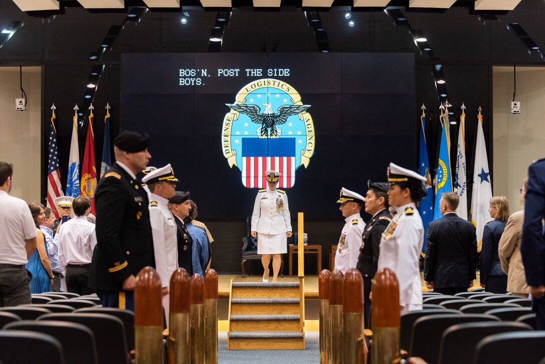 A woman in Navy dress whites (skirt version) with gold trim and many medals and ribbons stands after stepping up four steps in the middle of the stage. She is flanked by large bullet reproductions and military members from the Air Force, Navy and Army acting as her Sideboys.