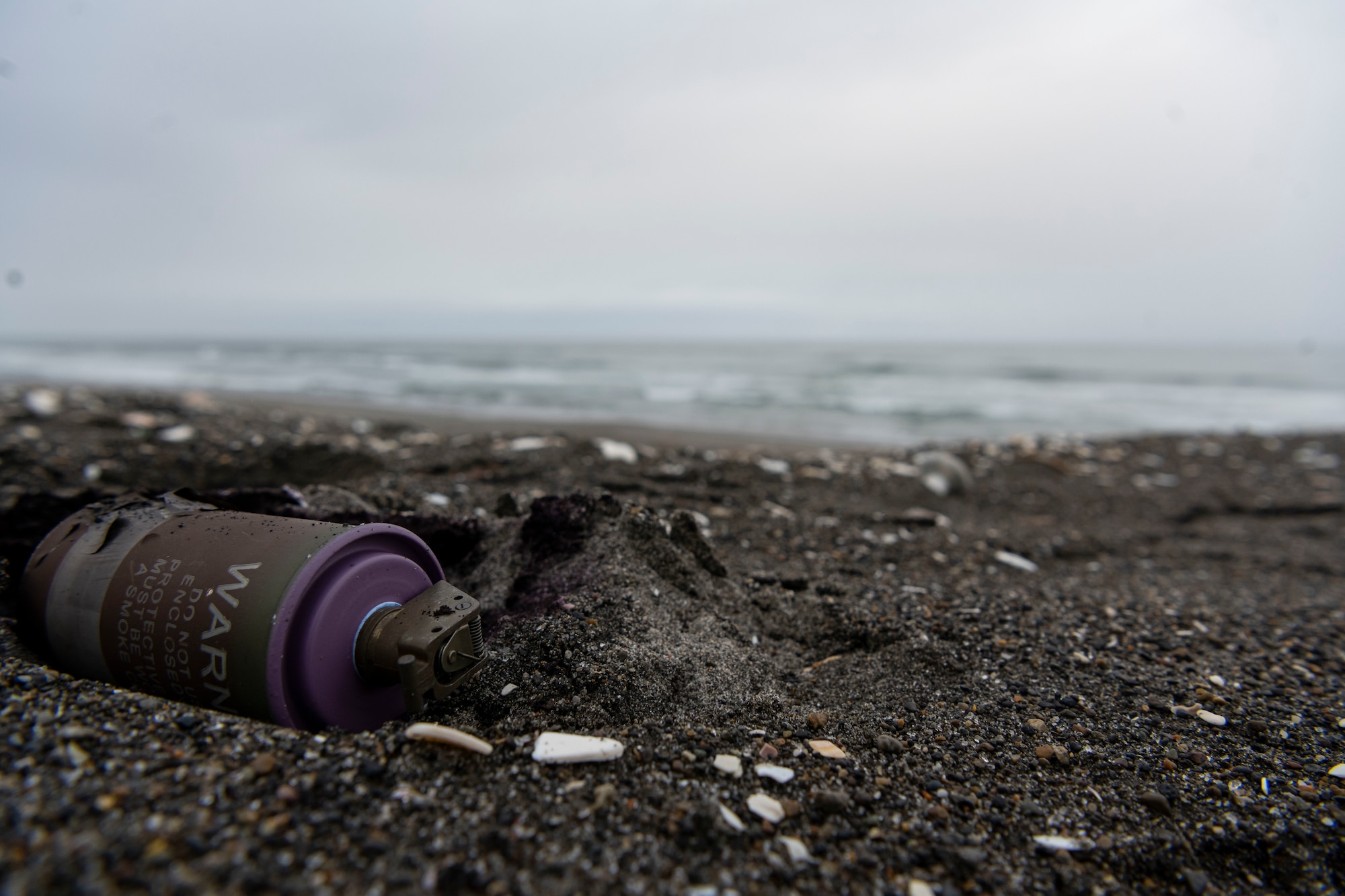 A purple smoke grenade lays in the sand of a beach.
