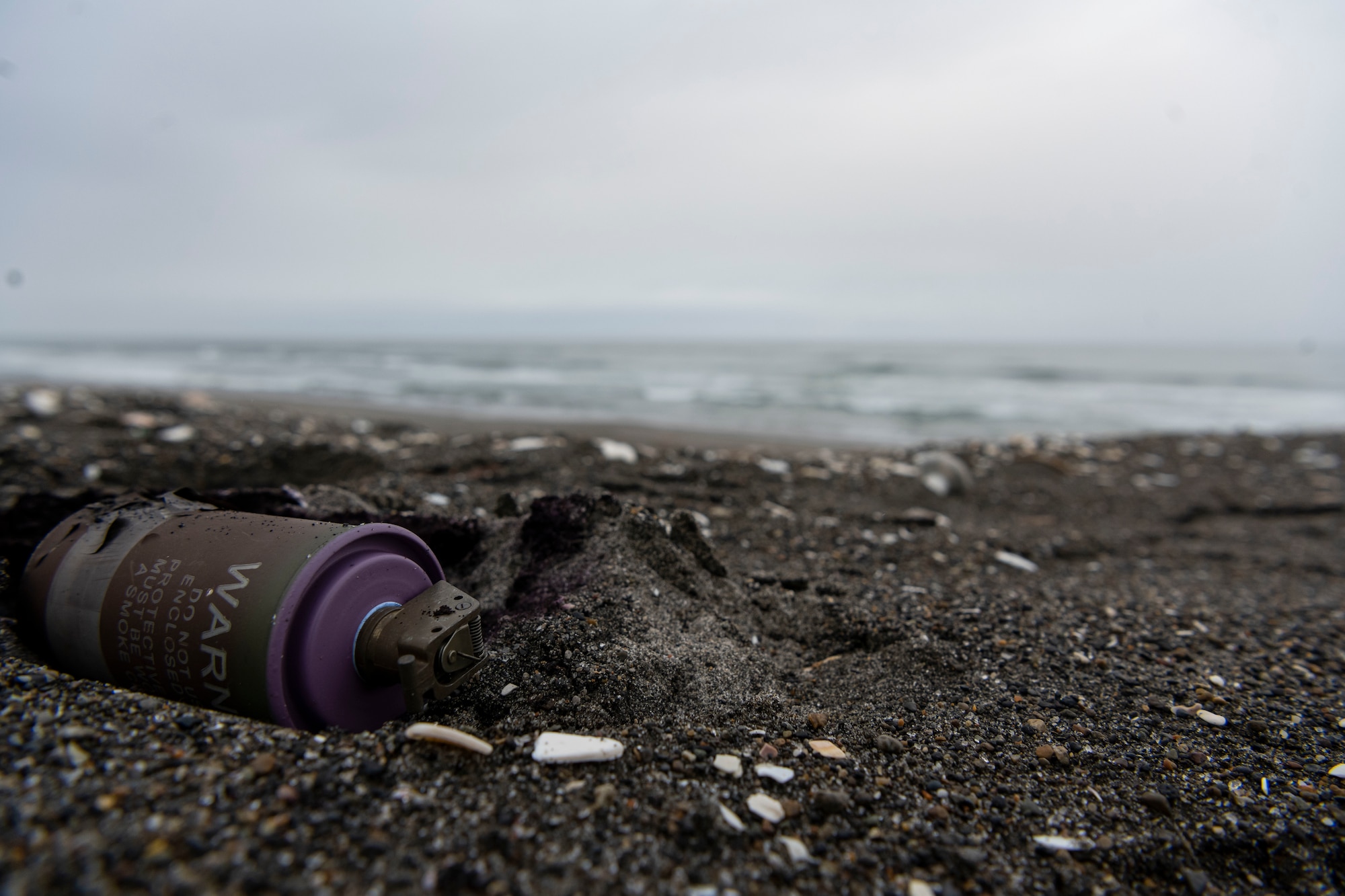 A purple smoke grenade lays in the sand of a beach.