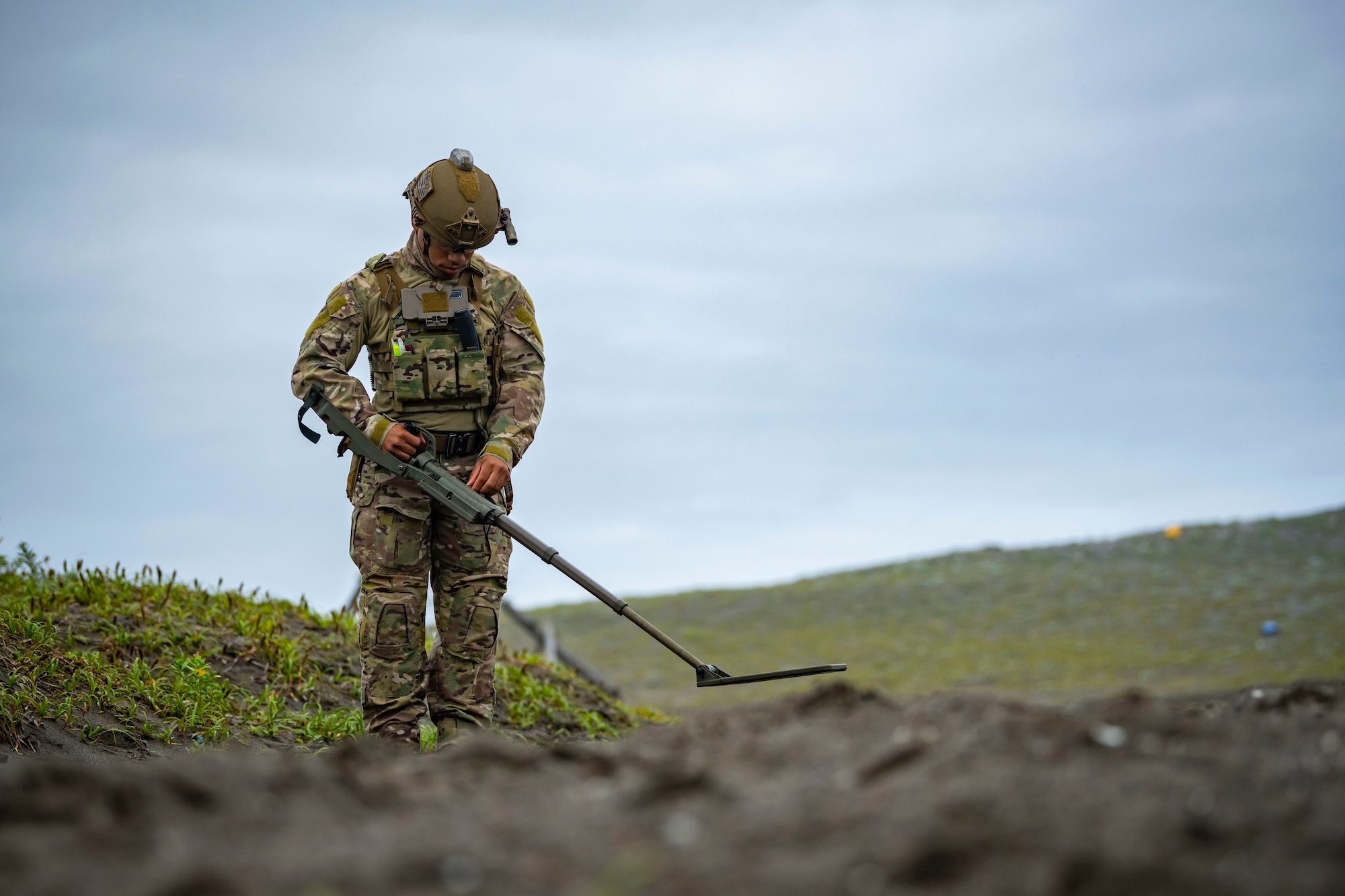 U.S. military member in tactical gear scans the ground with a metal detector.