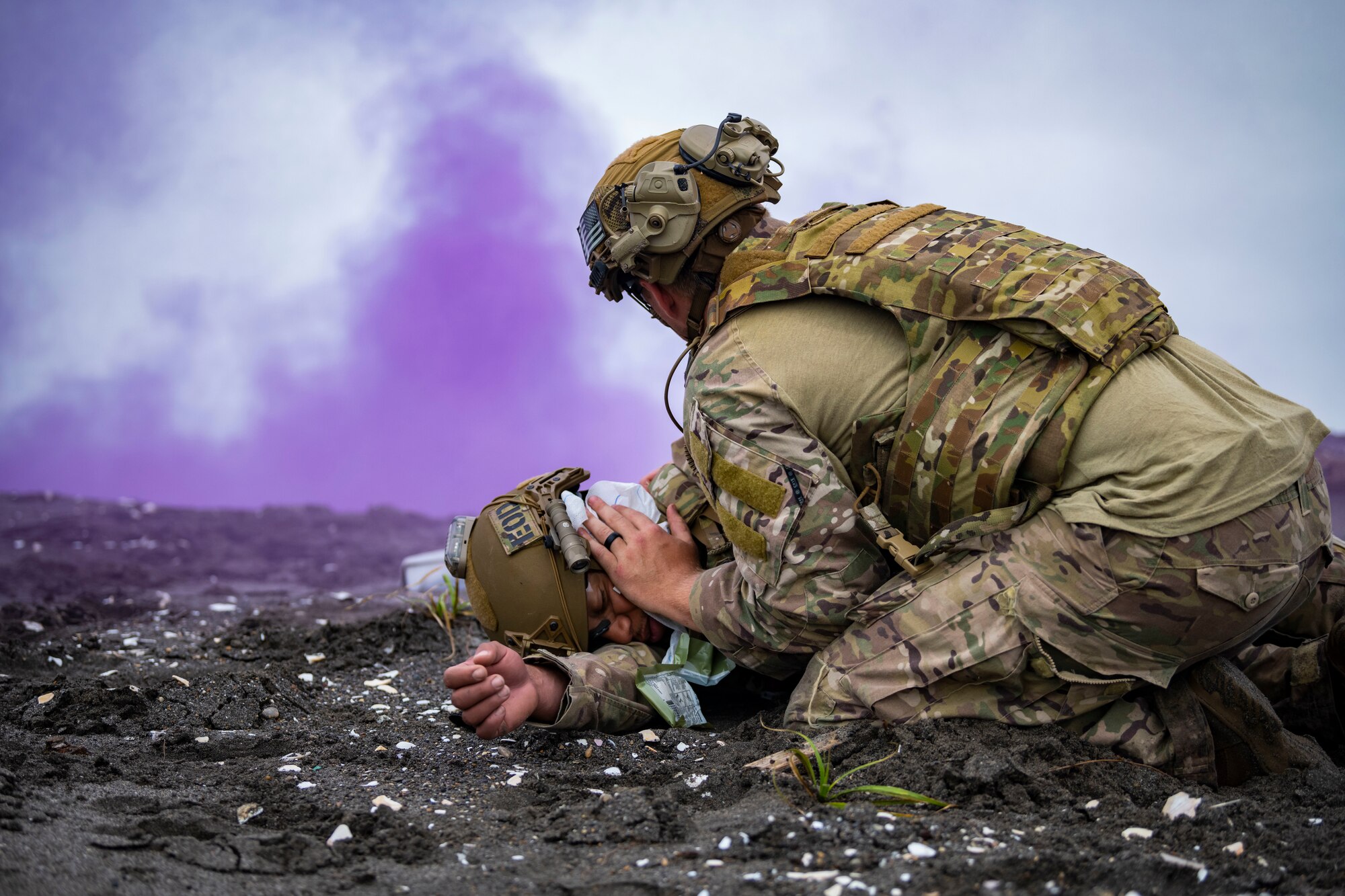 U.S. military members in tactical gear performs tactical combat casualty care as purple smoke spreads in the sky.