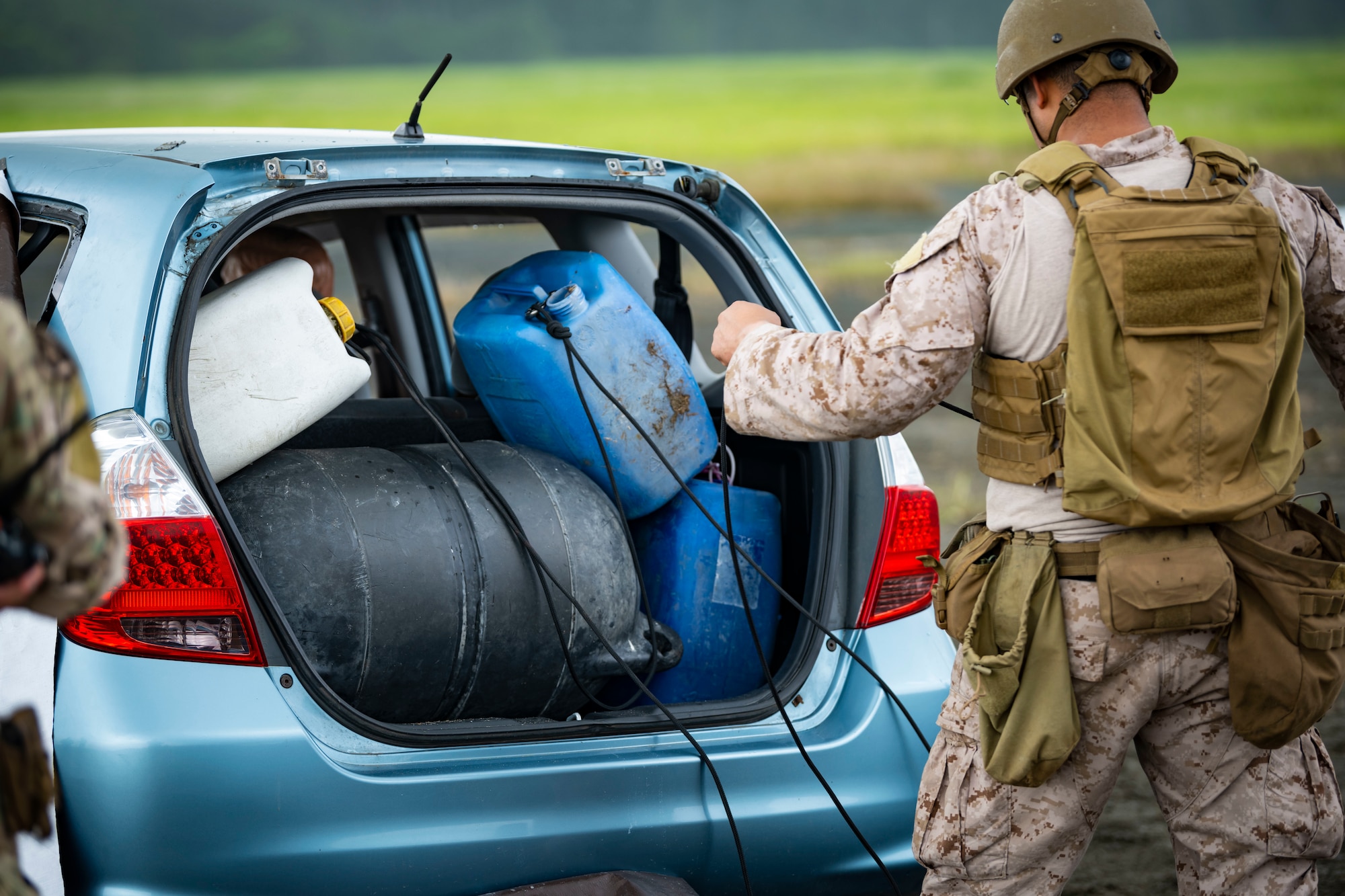 U.S. military member in tactical gear ties cables around multiple explosive ordnance in the trunk of a car.
