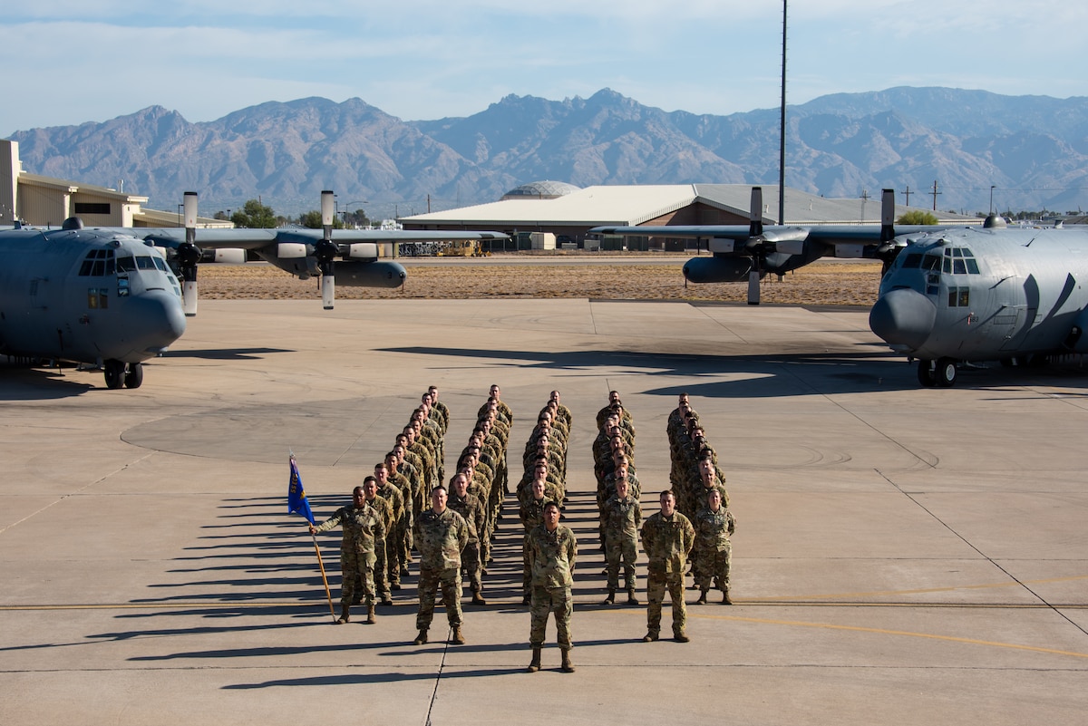 A group of uniformed Airmen stand in formation with the unit guidon for a photo in front of two EC-130H Compass Call aircraft.