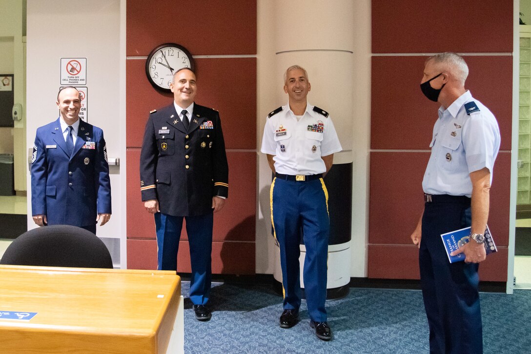 A man with a graying buzz cut wearing Air Force blues (short sleeve version) with no dress coat and wearing a black mask holds a program. He is chatting with two Army members in their dress uniforms and a Air Force Master Sergeant also in Air Force dress blues with his service coat on.