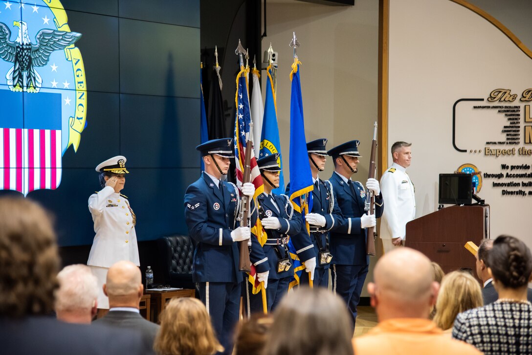 A woman in Navy dress whites salutes next to a color guard made up of Air Force members in dress blues with two holding rifles on the ends and one holding the U.S. Flag. To their right is a man with a graying buzz cut also in Navy dress whites. He is not saluting.
