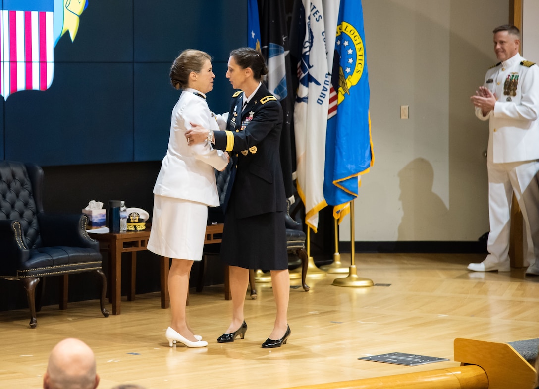 A woman with dark hair in a tight bun and wearing Army dress blues greets a woman in Navy dress whites on stage. The uniform is a dark almost black color and has gold trim. She wears a aviator badge and many ribbons and badges. The white uniform also has gold trim.