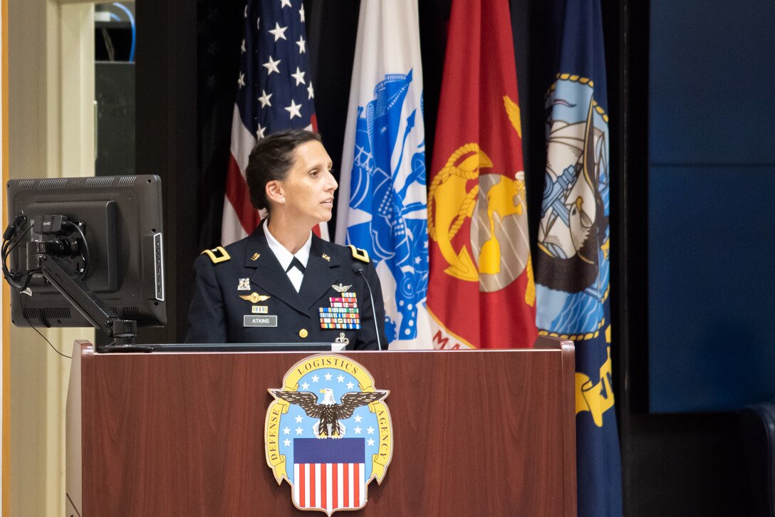 A woman with dark hair in a tight bun and wearing Army dress blues speaks at a podium in an auditorium. The uniform is a dark almost black color and has gold trim. She wears a aviator badge and many ribbons and badges.