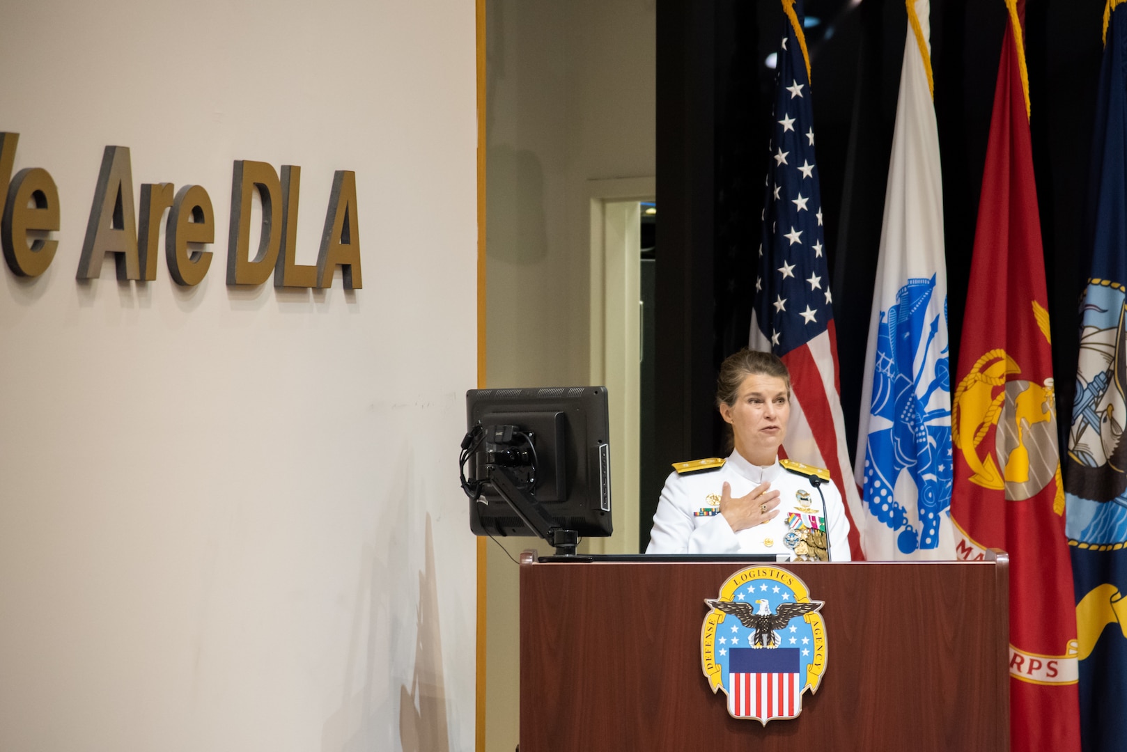 A woman with braided hair in a bun stands at a podium with a the US Flag and military flags in the background. She wears a pure white Navy uniform with gold trim and many medals and badges.