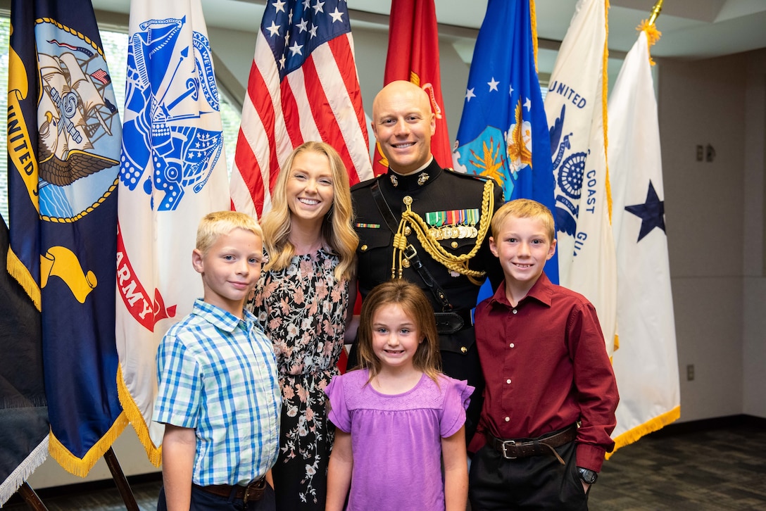 A man in a black and gold Marine Corps uniform with a blond woman and two boys and a girl with flags in the background.