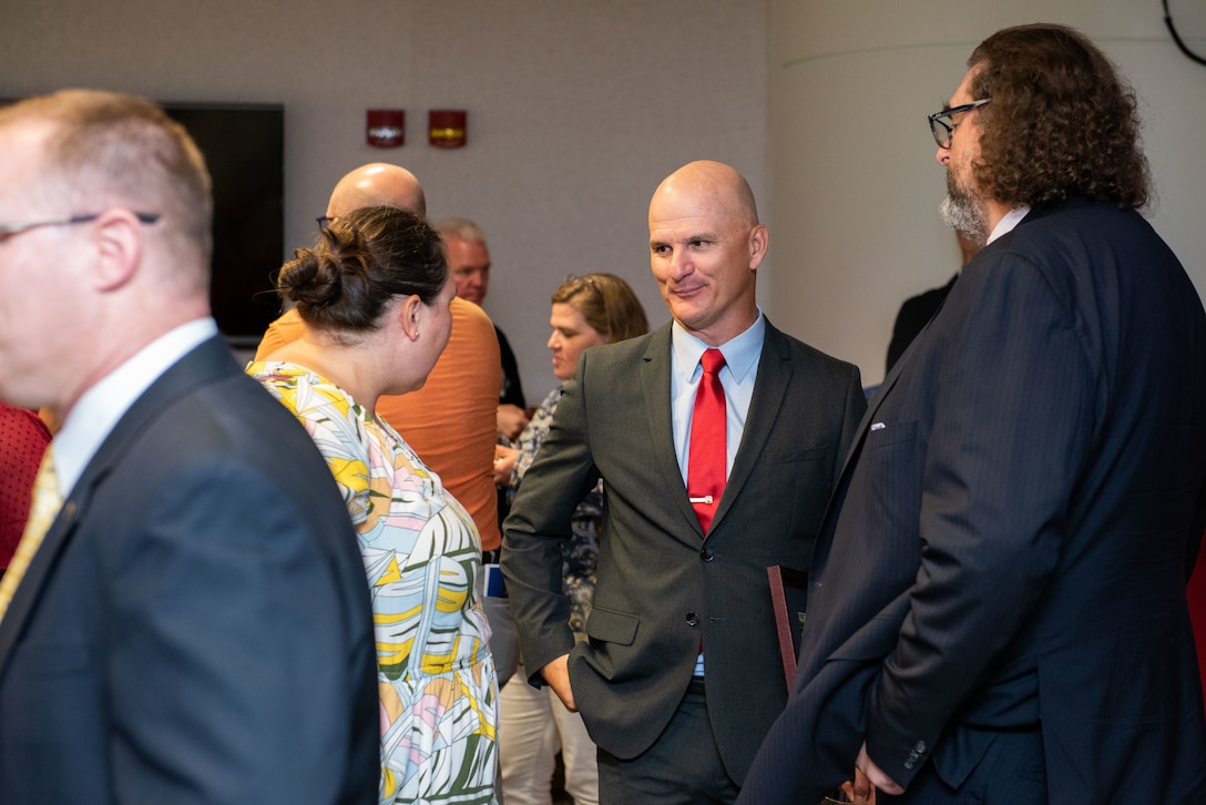A bald man in a dark suit chats with a man in a dark blue suit and a woman in a yellow flowered dress.