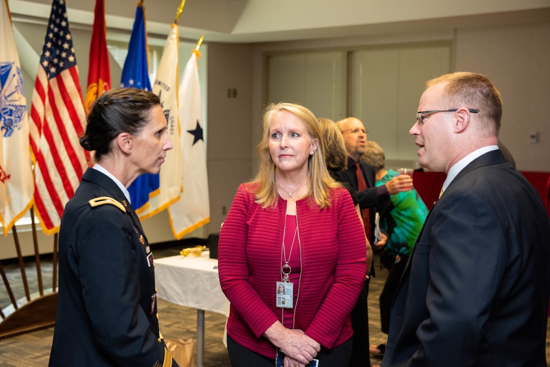 A woman in Army dress blues chats with a blond woman in a red blouse and a man in a grey suit with a buzz cut. The uniform is more black in color than blue and is trimmed with gold.