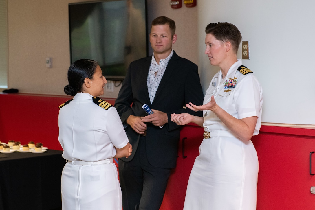 Attendees of a reception chat with one another. Two of the people are women in Navy dress whites and the third is a man in a dark suit.