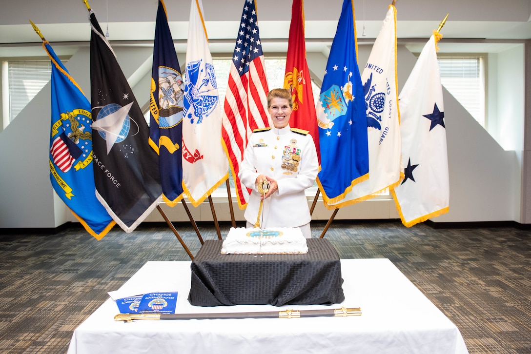 A woman holds a sword which will cut a square cake on a table with military flags behind her. She is in Navy dress whites with gold trim.