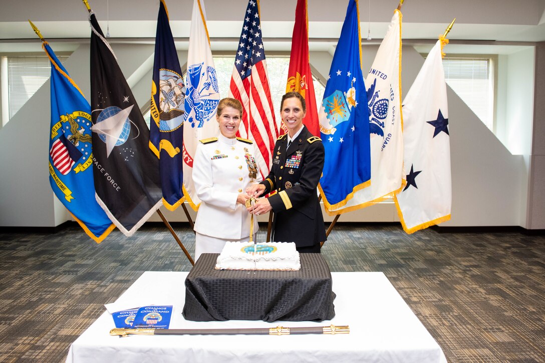 Two women hold a sword which will cut a square cake on a table with military flags behind them. One woman is in Navy dress whites and the other woman is in Army dress blues which are really black in color. Both uniforms have gold trim.
