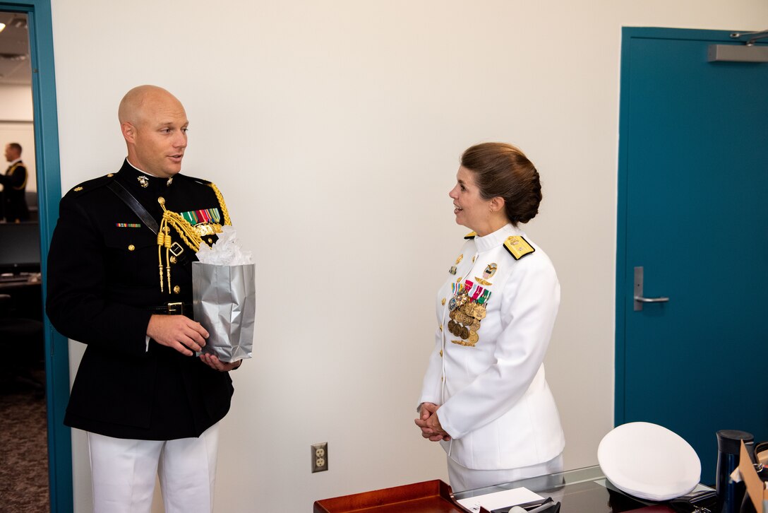 A man in a black and gold Marine Corps uniform  gives a woman in a pure white Navy uniform with gold trim smile a gift.