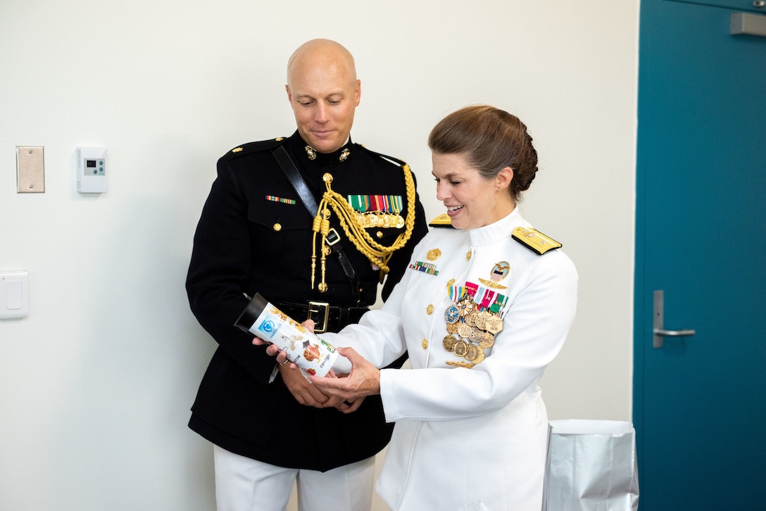 A man in a black and gold Marine Corps uniform with a woman in a pure white Navy uniform with gold trim smile as she opens a gift.