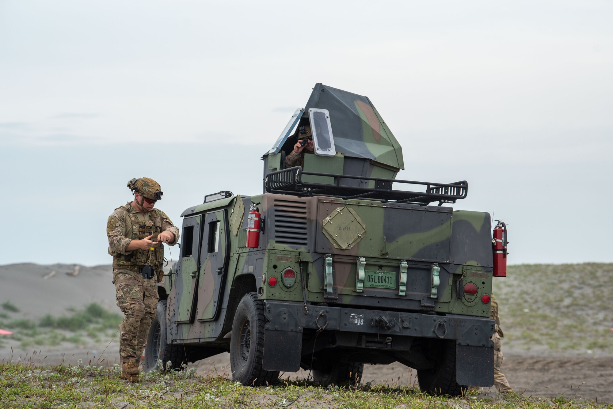 U.S. military members in tactical gear stands next too and looks out of a Humvee.
